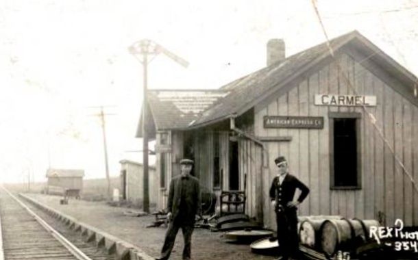 Black and white photo of the Carmel Depot along the Monon railroad with two railroad workers in uniform standing in front of the building alongside the railroad tracks. Photo courtesy of the Carmel Clay Historical Society.