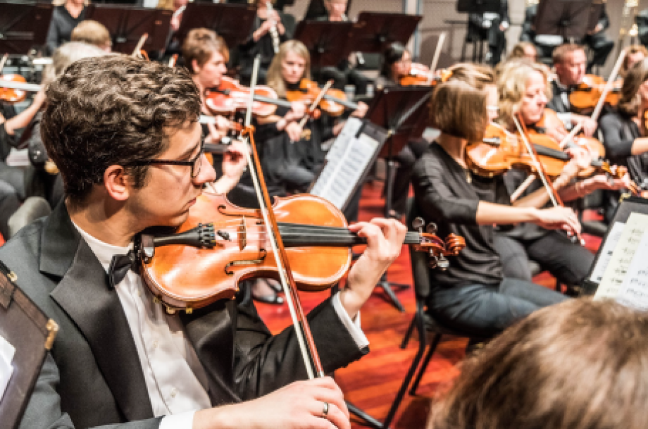 Close up on a male violinist in tuxedo at a performance with string section in the background on stage at the Payne & Mencias Palladium