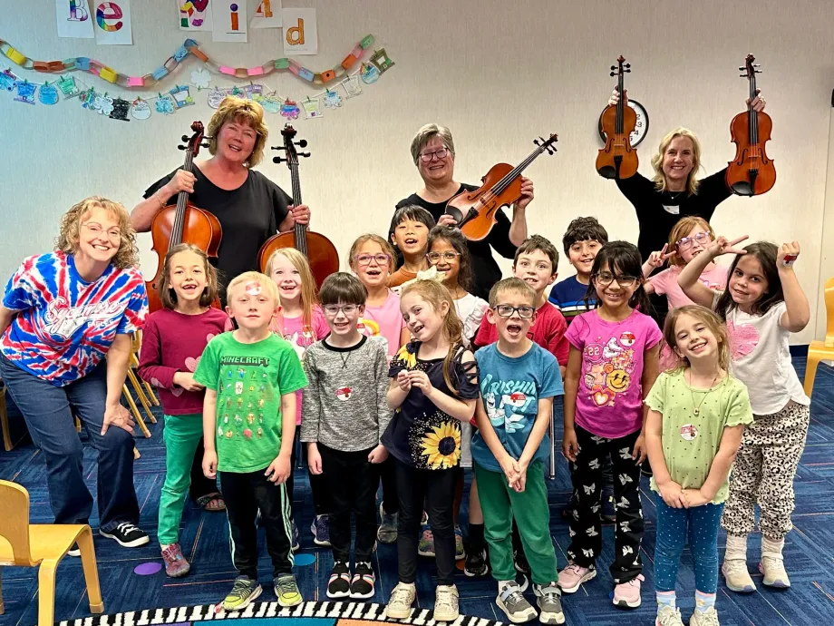 A group of elementary school students stand with three Carmel Symphony Orchestra musicians who are holding up half size string instruments used in an instrument "Pettin Zoo" where children can try to play these instruments to pique their interest in classical music and symphony orchestras