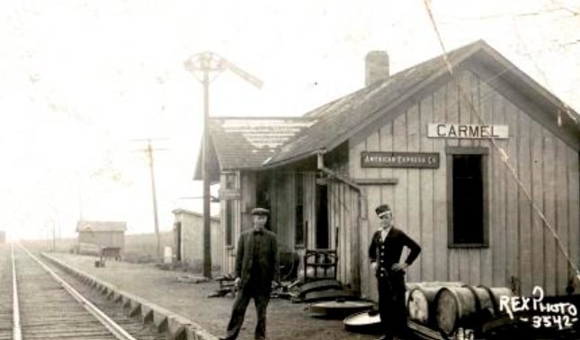 Black and white photo of the Carmel Depot along the Monon railroad with two railroad workers in uniform standing in front of the building alongside the railroad tracks. Photo courtesy of the Carmel Clay Historical Society.