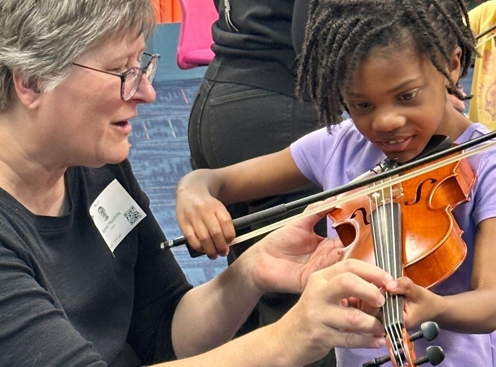 Young student trying out a smaller sized violin assisted by a Carmel Symphony Orchestra musician at an Instrument Petting Zoo event.
