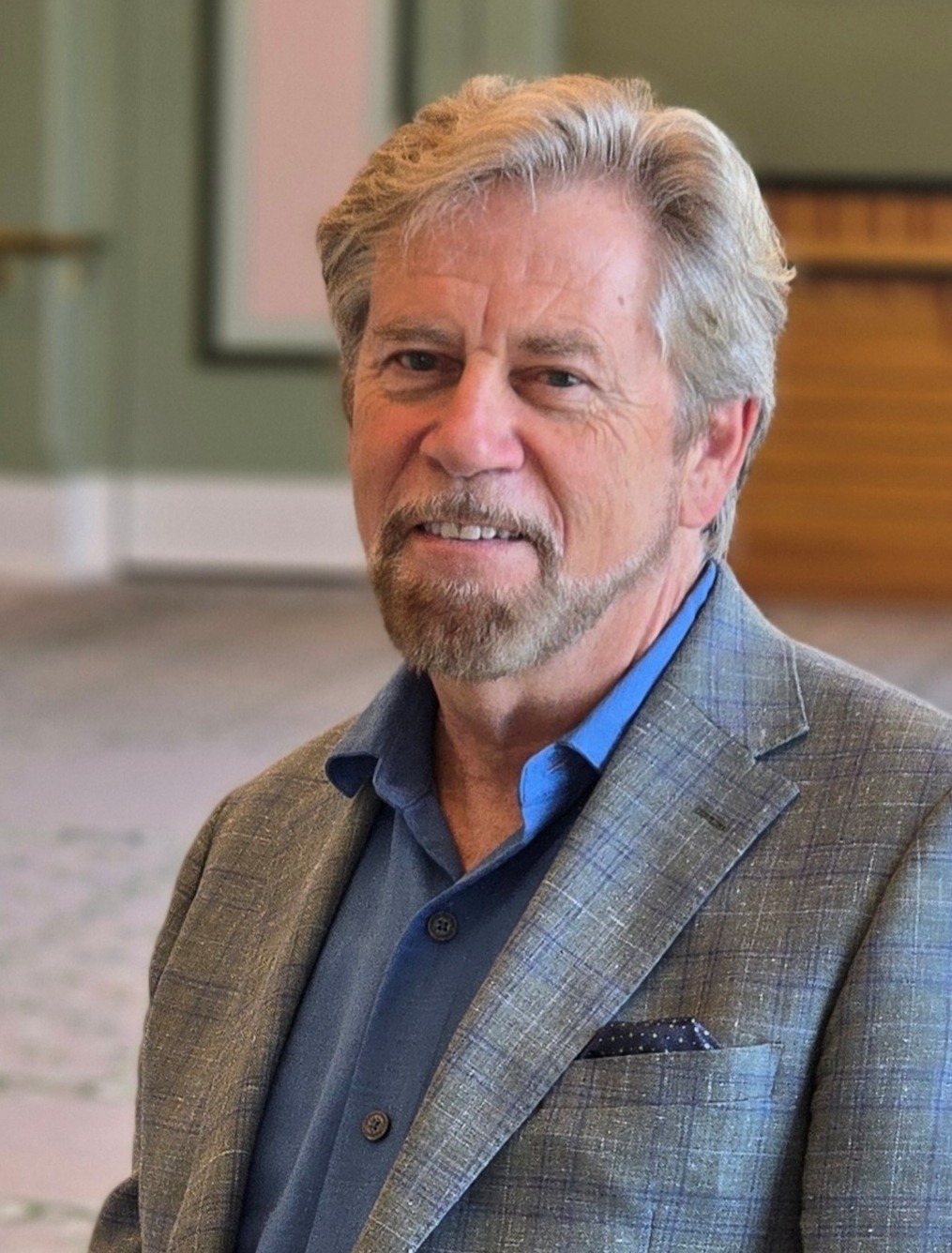 David Commanday, Music Director and Conductor of the Carmel Symphony Orchestra seated wearing a medium blue shirt and a plaid jacket in soft tones of blue and green