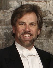 David Commanday, Music Director and Conductor for Carmel Symphony Orchestra in tuxedo with white bowtie standing in front of brick wall