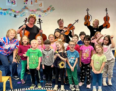 A group of elementary school students stand with three Carmel Symphony Orchestra musicians who are holding up half size string instruments used in an instrument "Pettin Zoo" where children can try to play these instruments to pique their interest in classical music and symphony orchestras