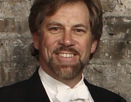 David Commanday, Music Director and Conductor for Carmel Symphony Orchestra in tuxedo with white bowtie standing in front of brick wall
