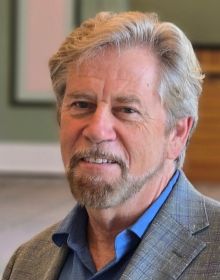David Commanday, Music Director and Conductor of the Carmel Symphony Orchestra seated wearing a medium blue shirt and a plaid jacket in soft tones of blue and green